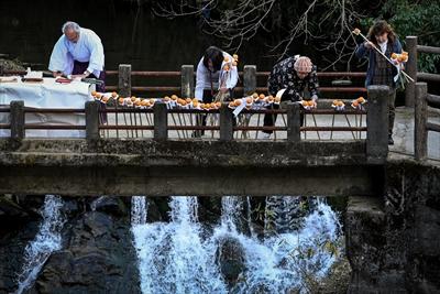 飛鳥川に架かる橋の上で綱掛神事の準備をする神主さんとミカンとお札をさした竹の棒をたくさん立てかけている女性三人の写真