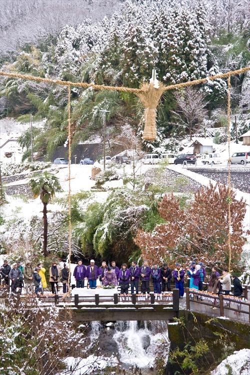雪が積もる中、完成したおづなと飛鳥川に架かるかんじょばしの上で祭事をする神主さんと参加者23人の写真